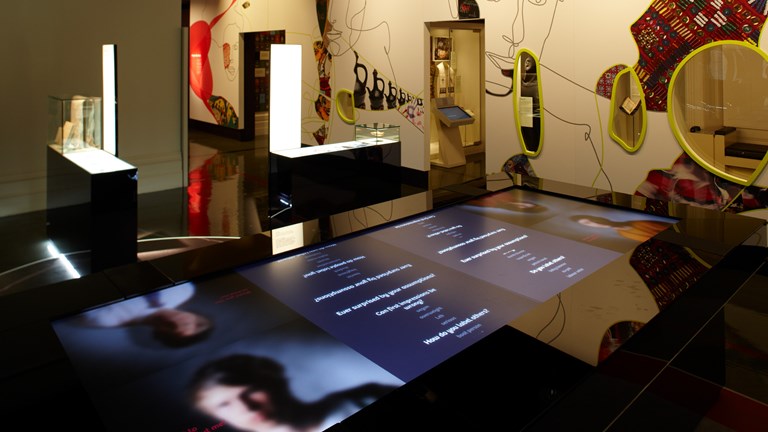A photo of Identity exhibition. There are video screens, objects in glass cases and walls with abstract art.