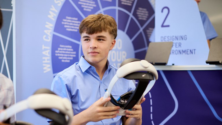 A high school student in a school uniform holds a virtual reality headset. He is sitting on a chair and is preparing to participate in the Road Trip to Zero, an interactive VR experience that is part of Road Smart Interactive, an incursion program that visits Victorian secondary schools.
