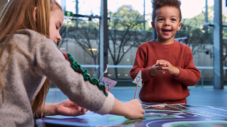 Two children have coloured in and cut out paper road signs that they’ve added to wooden sticks and placing on a vinyl road map.