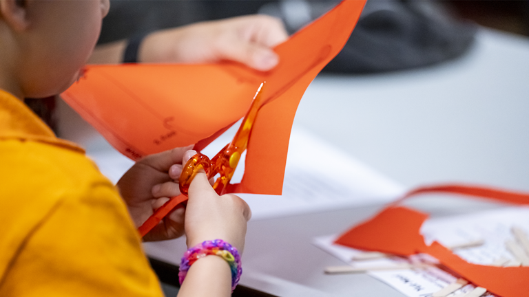 A close-up of a child cutting a red paper with plastic scissors, while an adult hand hold pulls the other side of the paper taut.